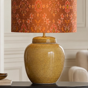 Könnte beinhalten: Eine Tischleuchte mit zylindrischem Lampenschirm in einem orange-roten Ikat-Muster. Der Fuß besteht aus einem bauchigen, senfgelben Keramikgefäß mit sichtbarer Craquelé-Glasur, das von einem kleinen goldfarbenen Metallhals gehalten wird.