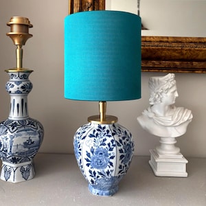 Pode incluir: Uma luminária de mesa de cerâmica azul e branca com uma cúpula turquesa. A luminária tem uma base de latão e um design floral. À esquerda, outro vaso de cerâmica azul e branco. Um busto branco à direita.