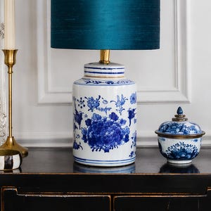 Op de afbeelding: Een keramische tafellamp met een teal lampenkap en een blauw-wit bloemmotief. De lampvoet heeft de vorm van een pot met een bijpassende kleine container met deksel. Links staat een gouden kandelaar.