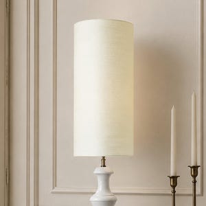 May include: A table lamp featuring a tall, cylindrical, off-white fabric lampshade with a subtle horizontal texture. The base is made of smooth, white ceramic with a curved, hourglass-like silhouette and a small metallic connector piece.