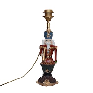 Puede incluir: Lámpara decorativa con diseño de soldado cascanueces. La lámpara tiene una parte superior dorada, un sombrero azul, una chaqueta roja y una barba blanca. La base es marrón con detalles dorados, y un cable dorado se extiende desde la lámpara.
