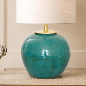 Puede incluir: Una lámpara de mesa con una base redonda de cerámica en color verde azulado con un acabado brillante y moteado. La lámpara incluye un cuello de metal dorado pulido y una pantalla cilíndrica de tela color blanco roto.