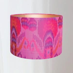 Op de afbeelding: Cilindrische lampenkap met een levendig ikatpatroon in roze, oranje en paarse tinten. De binnenkant van de lampenkap heeft een gouden afwerking, wat een warme gloed creëert. Een decoratief verlichtingselement.