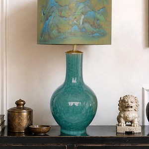 Può includere: Una lampada da tavolo con una base in ceramica lucida color verde acqua con finitura craquelé. Il paralume cilindrico mostra una scena paesaggistica stampata con montagne, alberi e figure in tonalità di verde, blu e oro.