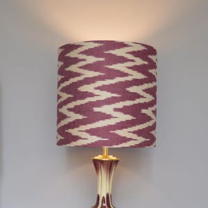 Op de afbeelding: Decoratieve tafellamp met een cilindrische lampenkap. De lampenkap heeft een zigzagpatroon in paars en crème. De lampvoet is crème, goud en bordeaux. De lamp is ingeschakeld en verlicht de kamer.