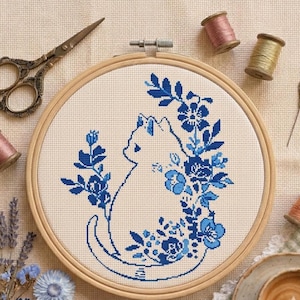 May include: A circular wooden embroidery hoop containing a cross-stitch pattern of a white cat surrounded by blue floral motifs. The project is accompanied by vintage-style metal scissors, several wooden spools of thread, and a single sewing needle.