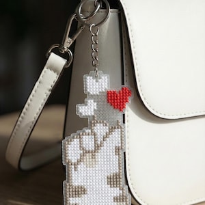 May include: A pixelated cat keychain with a red heart, attached to a silver chain and clasp. The cat is white and tan, made with a cross-stitch design. The keychain hangs from a white purse.