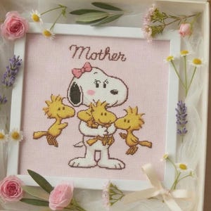 May include: Cross-stitch artwork featuring Snoopy and Woodstock with the word "Mother" above. The image is framed in white and surrounded by pink roses, daisies, and lavender sprigs, presented in a decorative box.