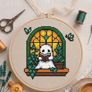May include: A cross-stitch project in a round wooden hoop featuring a white ghost reading a brown book inside a green arched window frame with yellow panes. The design includes green potted plants, two small green butterflies, and a brown ledge.