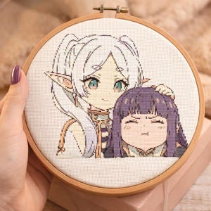 May include: A completed cross-stitch embroidery featuring two anime-style characters in a wooden hoop. One character has white hair and the other has purple hair. The artwork is on a white fabric background.