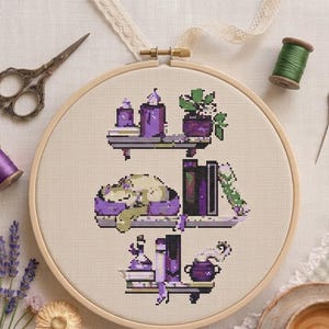 Peut inclure: Un projet de point de croix dans un tambour à broder en bois représentant trois étagères avec des bougies violettes, une plante en pot, un chat endormi, des livres, une fiole et un chaudron. Inclut des ciseaux vintage, du fil et une aiguille.