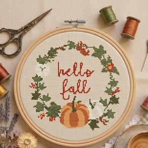 Può includere: Un telaio da ricamo rotondo in legno contenente un motivo a punto croce di una ghirlanda composta da foglie verdi, piccole bacche arancioni e una grande zucca arancione sul fondo. Il testo "hello fall" è ricamato in rosso al centro.