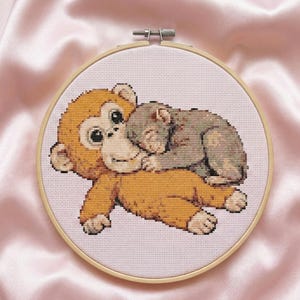 May include: Cross-stitch embroidery of two cartoon monkeys in a wooden hoop. One monkey is light orange, and the other is gray, with the gray monkey resting on the orange monkey. The background is light pink.