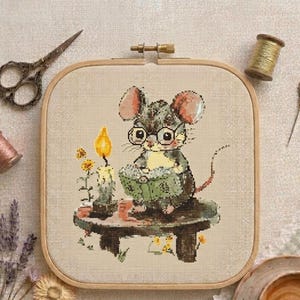 May include: A square wooden embroidery hoop containing a cross-stitch pattern of a grey mouse wearing round glasses and reading a green book. The mouse sits on a dark wooden stool next to a lit candle and small yellow flowers on a beige fabric.