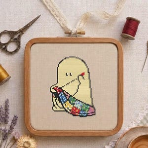Pode incluir: Um bastidor de bordado quadrado de madeira contendo um design em ponto cruz de um fantasma amarelo-pálido segurando uma colcha de retalhos colorida. O fantasma tem dois pequenos olhos pretos e um coração vermelho. Tesoura, linha e lavanda cercam o bastidor.