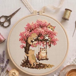 Op de afbeelding: Een ronde houten borduurring met een kruissteekpatroon van drie katten in een roze kersenbloesemboom. Eén kat zit op een schommel, één klimt in de stam en één kijkt uit de takken. Inclusief metalen schaar, garen en een naald.