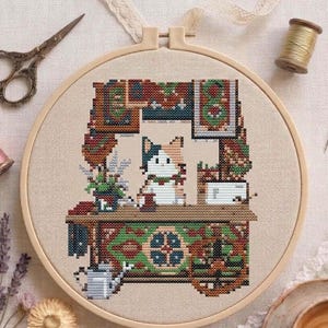 以下が含まれることがあります： 丸い木製の刺繍枠に入ったクロスステッチ刺繍作品。装飾された屋台の後ろに立つ三毛猫が描かれています。ベージュの布地に、緑と赤の模様のカウンター、小さなじょうろ、壁掛けのアートがデザインされています。
