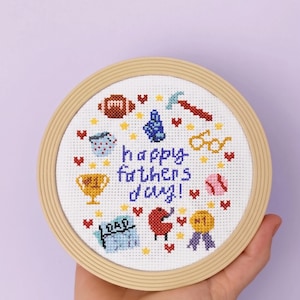 Könnte beinhalten: Runde Kreuzstich-Stickerei in einem Holzreifen. Das Design zeigt den Text "happy fathers day!" in Blau, umgeben von Symbolen wie Football, Hammer, Trophäe und Brille. Die Stickerei ist in einem farbenfrohen, handgemachten Stil gehalten.