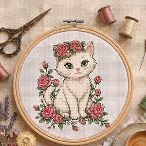 Può includere: Un telaio da ricamo circolare in legno contenente un motivo a punto croce di un gattino bianco con occhi verdi, che indossa una corona di rose rosa ed è circondato da cespugli di rose. Il telaio è circondato da forbici, rocchetti e un ago.