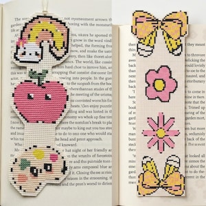May include: Two cross-stitch bookmarks. The left bookmark features a rainbow, a pink apple, and a paint palette. The right bookmark features two yellow bows, a pink flower, and a daisy, all stitched on a light cream fabric background.
