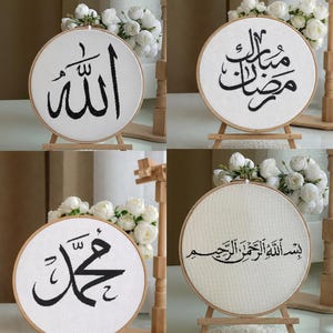 May include: Four round embroidery hoops with black Arabic calligraphy on white fabric. Each hoop is displayed on a small wooden easel. White flowers and wooden decorative elements are in the background.