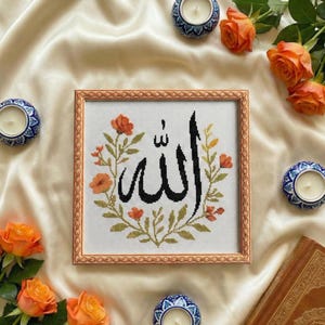 May include: A framed cross-stitch artwork featuring the Arabic word "Allah" in black script, surrounded by orange and green floral embroidery. The frame is a light brown color. The image is set against a cream-colored satin background, with orange roses and candles.