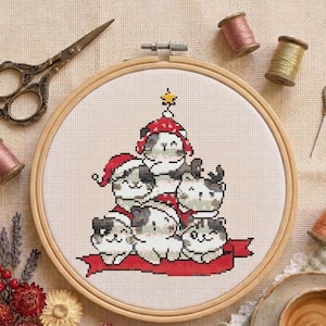 May include: A round wooden embroidery hoop containing a cross-stitch pattern of six stacked cats wearing red Santa hats and scarves, topped with a yellow star. The design sits on white fabric, surrounded by vintage scissors, thread spools, and a needle.