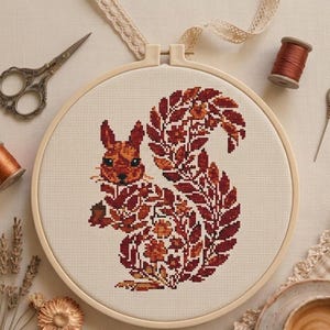 May include: A circular embroidery hoop containing a cross-stitch pattern of a squirrel composed of autumn-colored leaves and flowers in shades of red, orange, and brown on a cream-colored fabric, accompanied by sewing scissors, thread, and a needle.