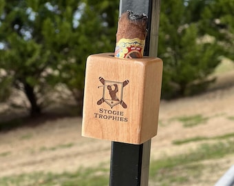 May include: Two wooden cigar holders with the text "STOGIE TROPHIES" and a golf-themed logo. One holder contains a lit cigar. The holders are attached to a black metal pole, with a blurred green background.
