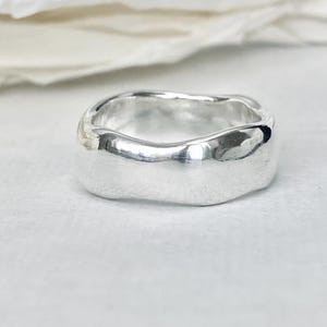 Peut inclure: Une bague argentée avec un anneau épais et irrégulier. La bague a une surface lisse et polie et un design légèrement ondulé. La bague est présentée sur une surface blanche.