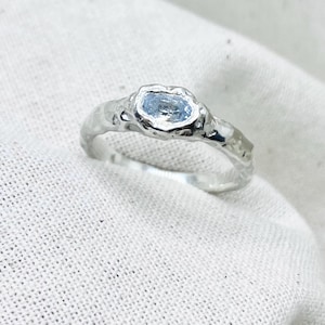 Könnte beinhalten: Ein silberner Ring mit einem hellblauen, oval geformten Edelstein. Das Band hat ein strukturiertes, organisches Design. Der Ring ist vor einem weißen, strukturierten Stoffhintergrund platziert. Der Edelstein ist der Mittelpunkt des Rings.