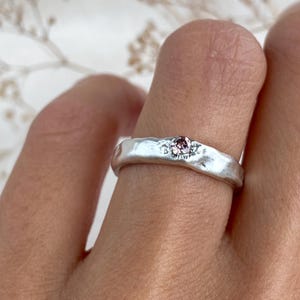 Könnte beinhalten: Ein silberner Ring mit einer texturierten, unebenen Oberfläche und einem einzelnen kleinen, runden, rosafarbenen Edelstein, der in die Mitte des Bandes eingefasst ist. Das Metall hat ein mattes, organisches Finish mit sichtbaren Vertiefungen.
