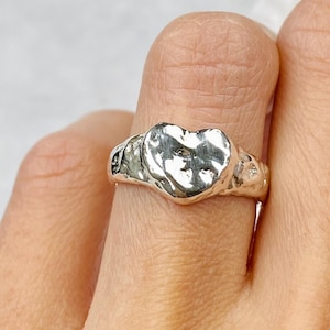May include: A silver ring with a textured, molten design. The ring has a heart-shaped centrepiece. The ring is worn on a finger, showcasing its unique, handcrafted appearance. The ring's surface reflects light, highlighting its metallic sheen.