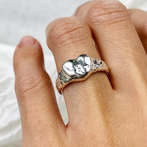 Könnte beinhalten: Ein silberner Ring mit einem strukturierten, organischen Design. Der Ring hat ein herzförmiges Mittelstück mit unebener Oberfläche. Das Band ist glatt und einfach und steht im Kontrast zur detaillierten Oberseite. Der Ring wird am Finger getragen.