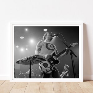 May include: Black and white photograph of a live music performance. A musician is playing an electric guitar with a unique design, while singing into a microphone. A keyboard is in front of the musician. Another musician is visible in the background.