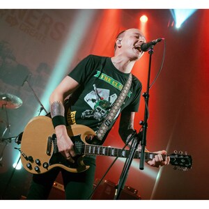 Puede incluir: Actuación en directo de un músico tocando una guitarra eléctrica marrón claro y cantando a un micrófono. El músico lleva una camiseta negra con un gráfico y una correa de guitarra estampada. El escenario está iluminado con luces rojas y blancas.