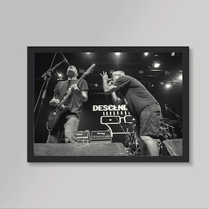 May include: Black and white photograph of a live music performance. Two people are on stage, one playing an electric guitar and the other singing into a microphone. The band's name, DESCENDENTS, is visible on the backdrop.