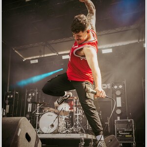 Puede incluir: Un hombre con un tanque rojo y pantalones negros está cantando en un micrófono en el escenario. Está sosteniendo el micrófono en su mano derecha y su brazo izquierdo está levantado en el aire. Está de pie frente a una batería y un altavoz.