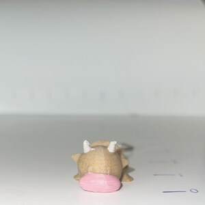 May include: A small, tan-colored cow figurine with white horns and a pink snout. The cow is lying down, with its head facing the viewer. The figurine is likely made of a hard material.