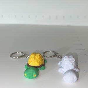 May include: Two turtle keychains. One keychain features a turtle with a yellow shell, green body, and brown trim. The other keychain is white. Both keychains have silver keyrings. The keychains are on a white surface.
