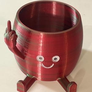 May include: A red and green 3D-printed planter with a cartoon face and legs. The planter has a hand giving the middle finger. The planter has a cylindrical shape with a wide opening.
