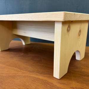 May include: A small, unfinished wooden step stool. The stool is rectangular with four legs and a curved cutout design. The wood has a light, natural color, and the stool measures approximately 18 inches long, 9 inches wide, and 8 inches high.