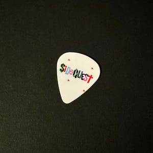 Puede incluir: Una púa de guitarra blanca con la palabra "SideQuest" impresa en letras coloridas. Pequeñas formas de estrella roja están dispersas alrededor del texto. La púa está sobre un fondo negro.