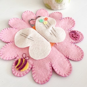 May include: A pink felt flower-shaped pincushion with a white felt centre. The pincushion has a bee and ladybug appliqué. There is a small pair of scissors and a white felt pincushion with green polka dots in the background.