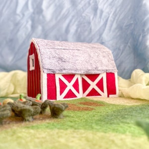 Puede incluir: Un granero de fieltro rojo con un techo gris y detalles en forma de X blancos en las puertas. El granero está sentado en un campo de fieltro verde con acentos de fieltro marrón. Hay varios pollos pequeños de fieltro marrón frente al granero.