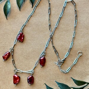 May include: A silver-toned chain necklace with five dark red, irregularly shaped beads. The beads are suspended from the chain at intervals. The necklace is displayed on a neutral background with green leaf accents.