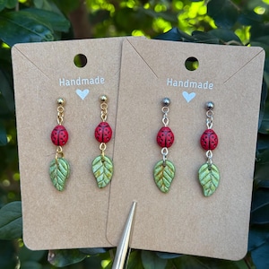 May include: Handmade earrings featuring a red ladybug design with black spots, a green leaf, and gold-colored accents. The earrings are displayed on a brown card with the word "Handmade" and a heart symbol.