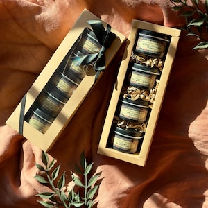 May include: Gift set of small, black tins with labels in a rectangular cardboard box. The box is tied with a dark green ribbon. The tins are filled with a product, and the box is filled with brown packing material.