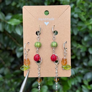 May include: Two pairs of silver-tone hoop earrings displayed on a brown card labelled Handmade. One pair features green ribbed beads, red ladybug charms, and dangling amber and green crystals. The second pair features amber tulip beads and green leaf charms.