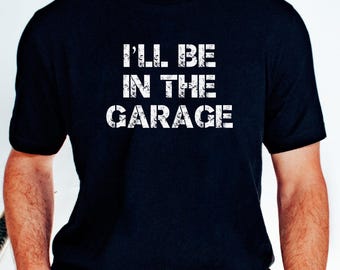 May include: Black t-shirt with the text "I'LL BE IN THE GARAGE" in distressed white lettering. The shirt is a crew neck style and is being worn by a person. The person is wearing blue jeans and a brown watch.